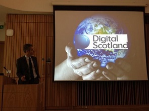 Speaker at a podium presenting beside a large screen showing a “Digital Scotland” slide.